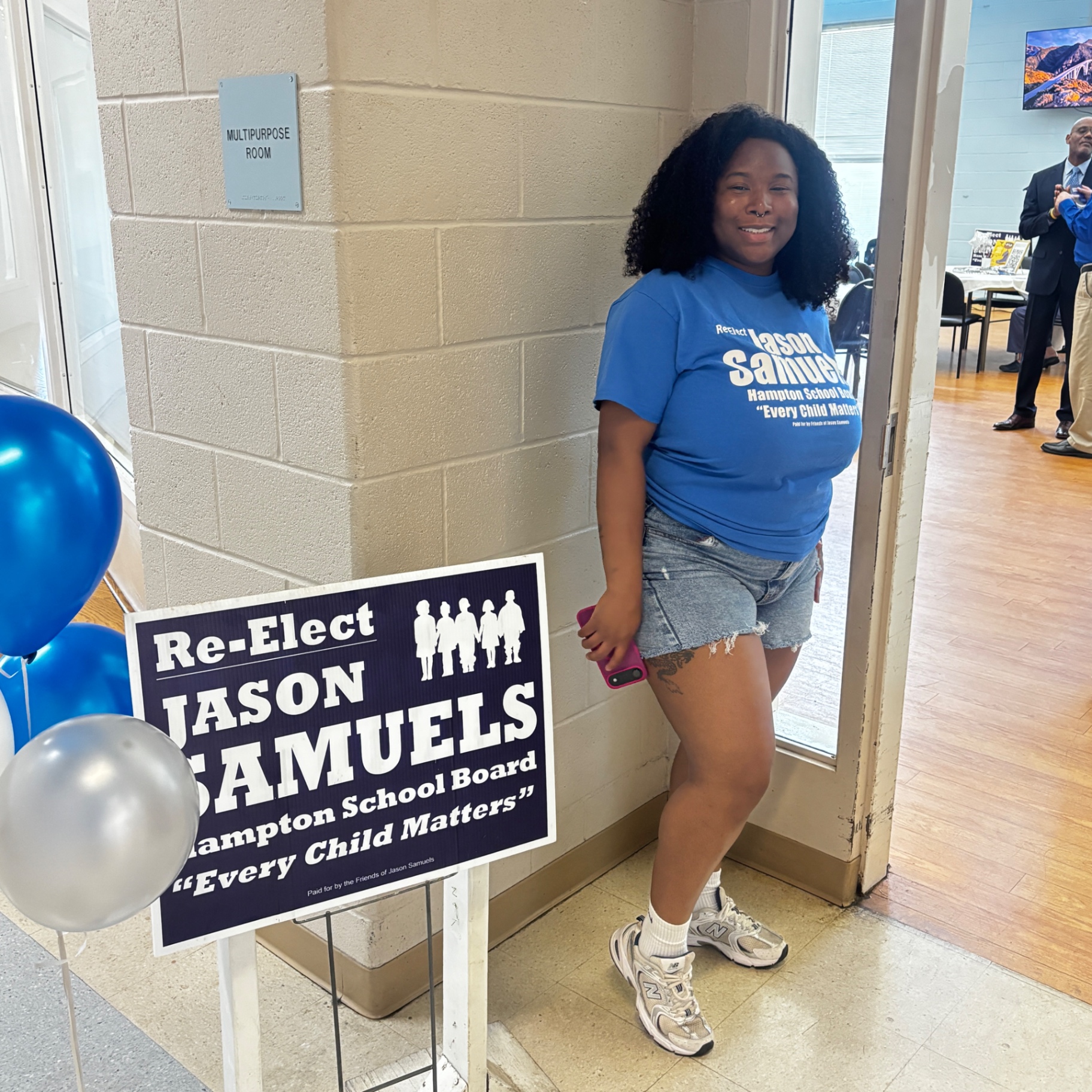 A volunteer in a Re-Elect Jason Samuels shirt poses beside a campaign sign reading Every Child Matters at the kickoff event.